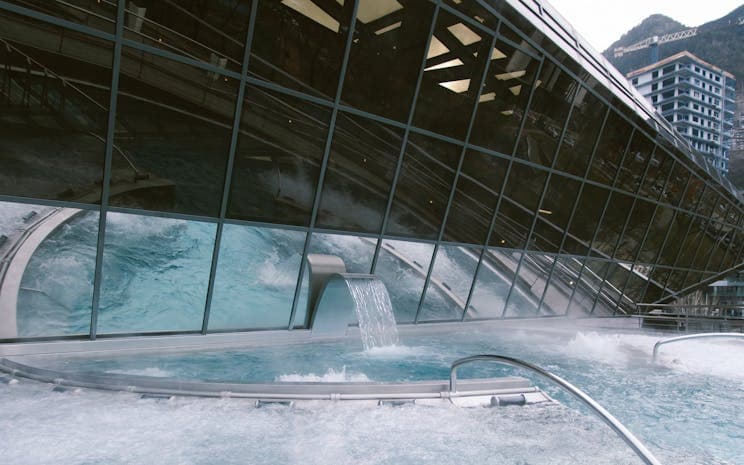 Exterior view of the Caldea thermal spa complex in Les Escaldes, featuring a large glass facade, a water feature, and surrounding mountainous architecture.