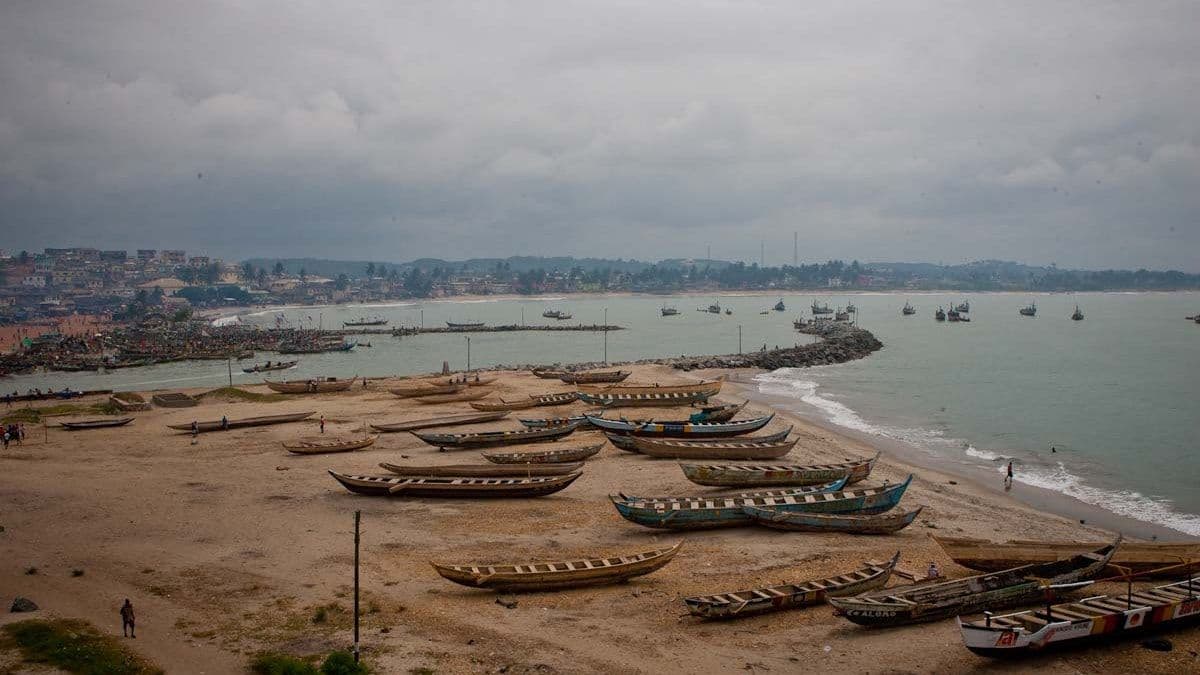 Coastal beach scene in Ghana's Volta Region with wooden fishing boats on sand, calm sea, and a distant shoreline town.