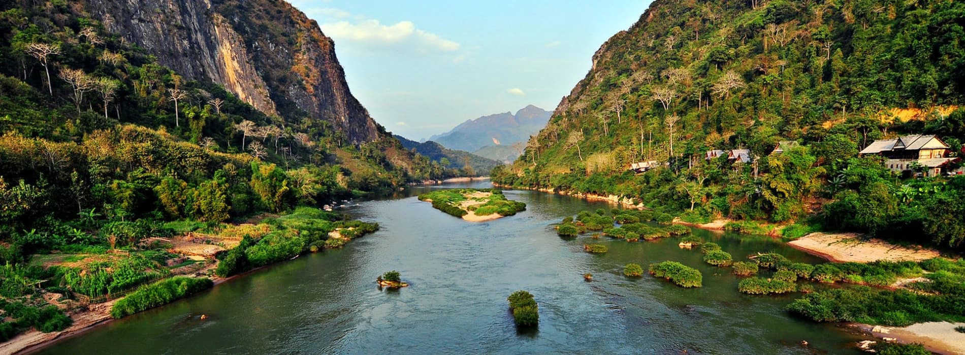 A wide river flows through a lush, forested valley flanked by steep limestone mountains and scattered rural houses.