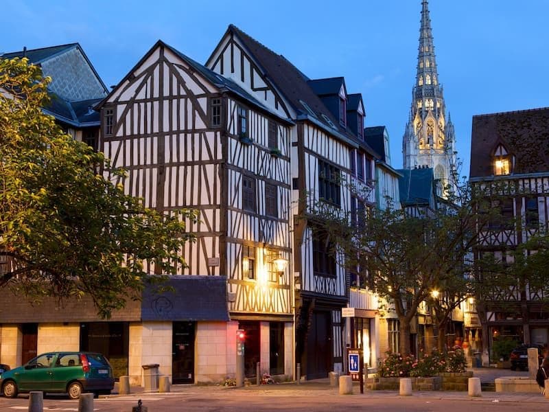 Half-timbered Normandy street at dusk with a church spire, parked car, and shopfronts.