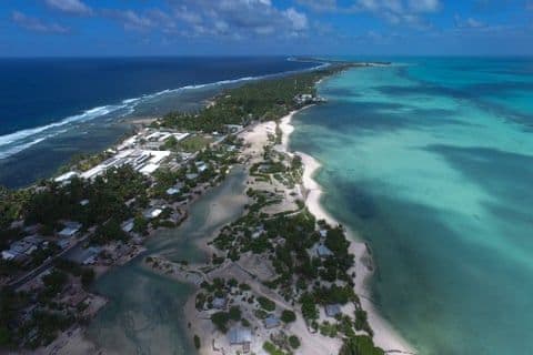 Aerial view of a narrow coastal village with turquoise ocean, white sand, and scattered buildings under a partly cloudy sky.
