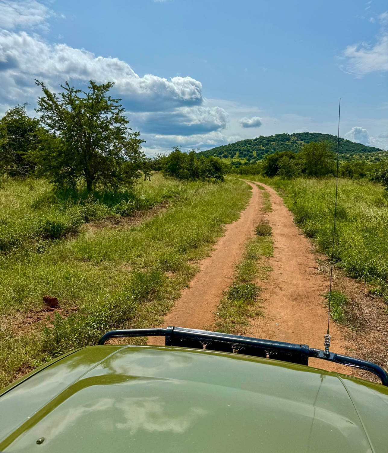 Dirt road through grassy savanna with green hills, viewed from a safari vehicle hood under a partly cloudy sky in the Kagera region.