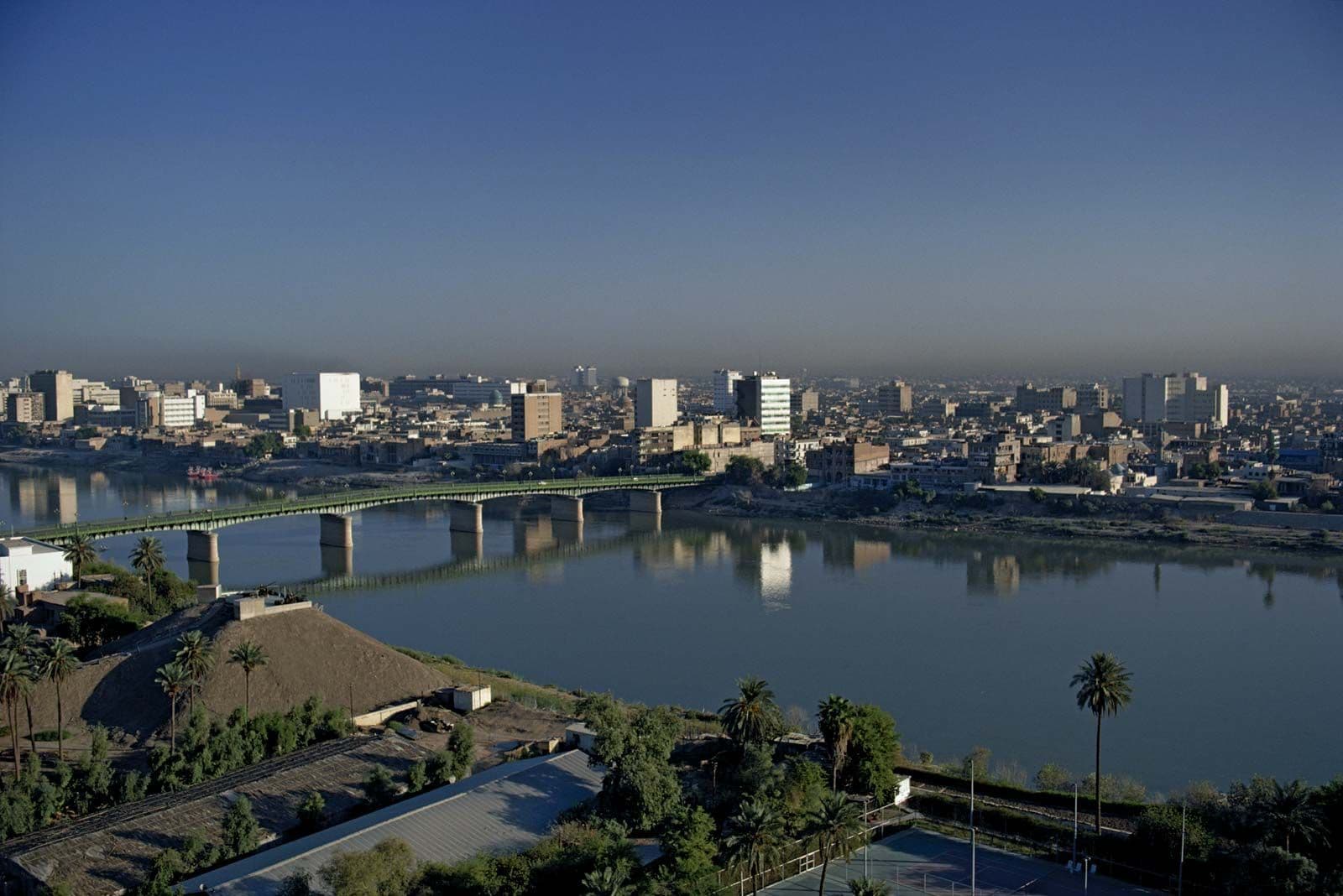 Wide view of the Tigris River in Iraq with a bridge, city skyline, and palm trees under a clear sky.