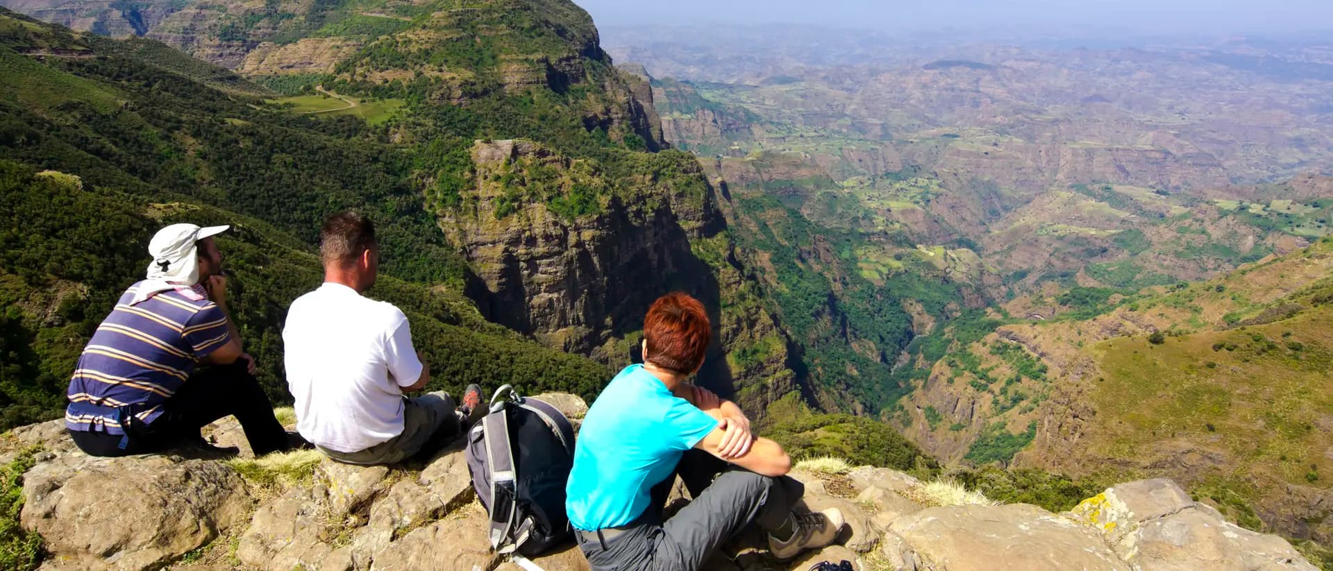 Three hikers sit on a rocky highland overlook, facing a vast dry-season valley landscape with distant ridges.