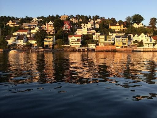 A scenic view of colorful houses lining a hillside along a fjord in Drøbak, with golden sunlight reflecting on the rippling water surface.