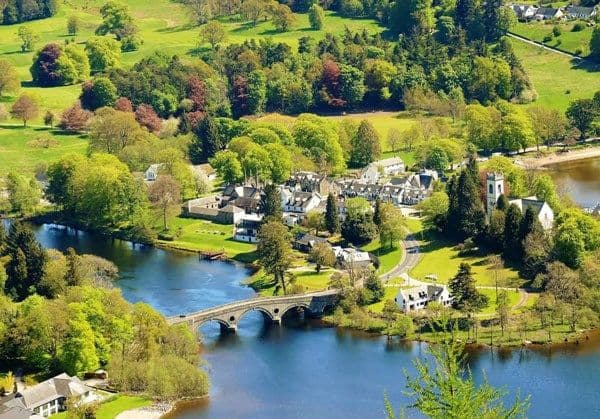 Aerial view of a calm river winding through a green village with a stone bridge and lush summer banks in the Perth region.