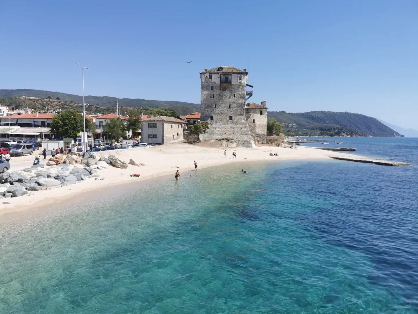 A scenic coastal view of Áfytos featuring a historic stone tower on a sandy beach, with clear turquoise water and hills in the background.