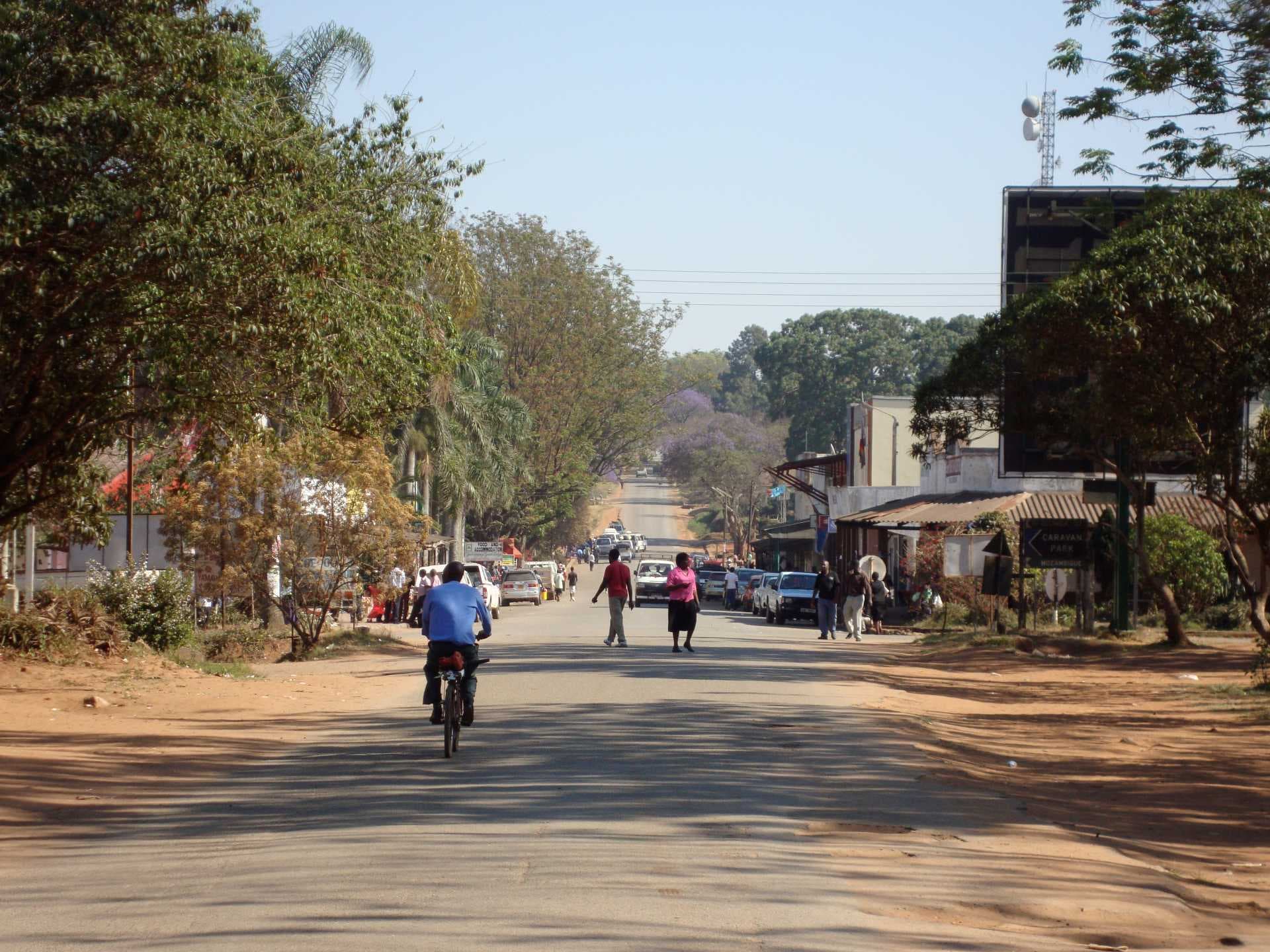 Street scene in Chipinge with a cyclist, pedestrians, parked vehicles, roadside shops, and trees under a clear sky.