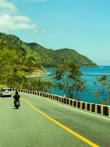 Paved coastal road with yellow center line, motorcycle and car, green hills, and blue sea under a clear sky