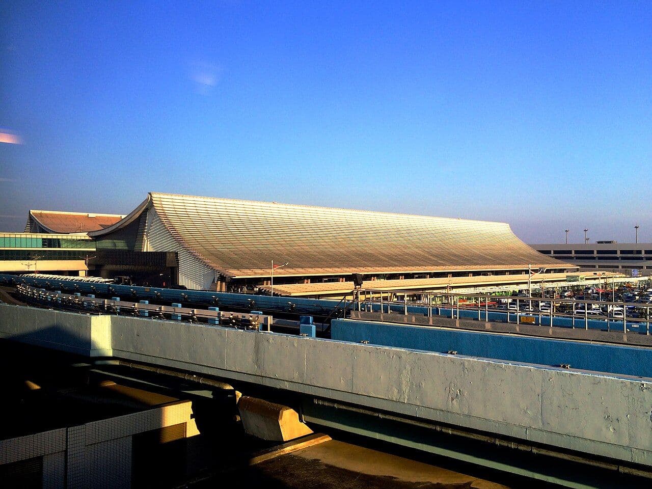 Exterior view of a modern transit hub with a large sloped roof, elevated rail tracks, and parking area under a clear blue sky.