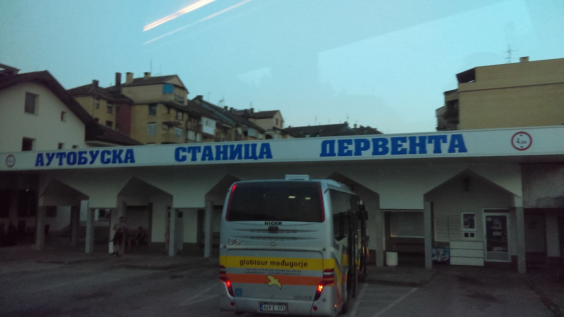 Bus parked at Derventa bus station with Cyrillic signage, residential buildings behind, and a few people in the background.