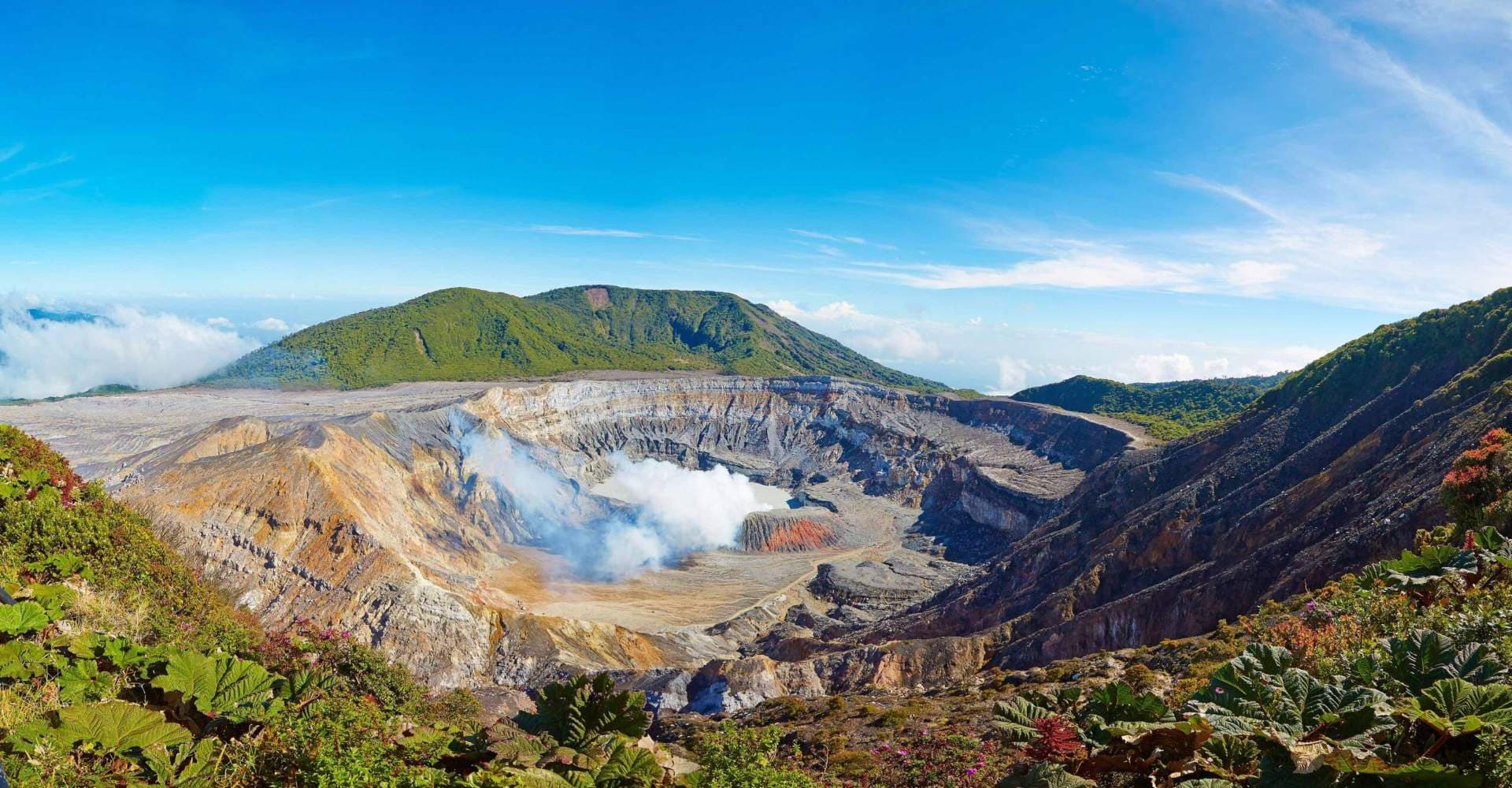 Panoramic view of Poás Volcano crater with steaming acidic lake, barren slopes, and surrounding green vegetation under blue sky.