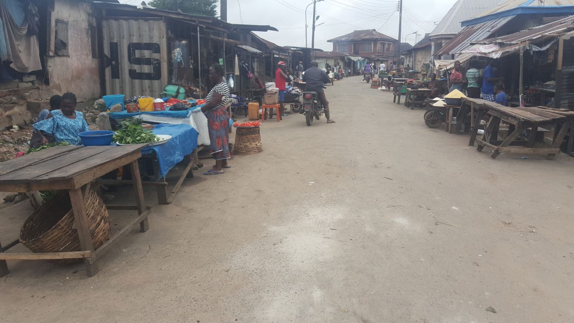 Busy outdoor market street scene with vendors selling produce on wooden tables and a motorcyclist passing by.
