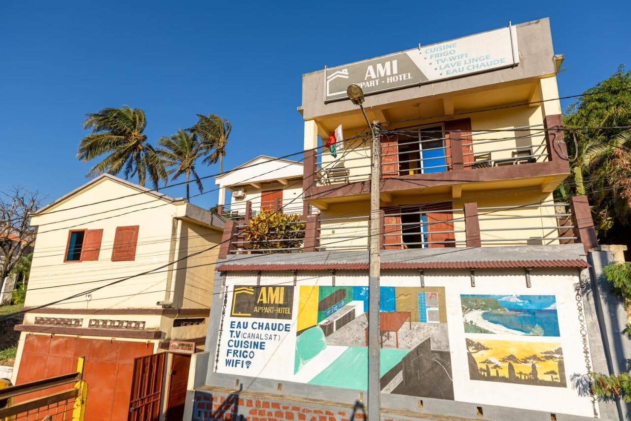 Exterior view of the Ami Appart-Hotel in Mahanoro, featuring a yellow building with balconies, palm trees, and a painted wall under a clear blue sky.