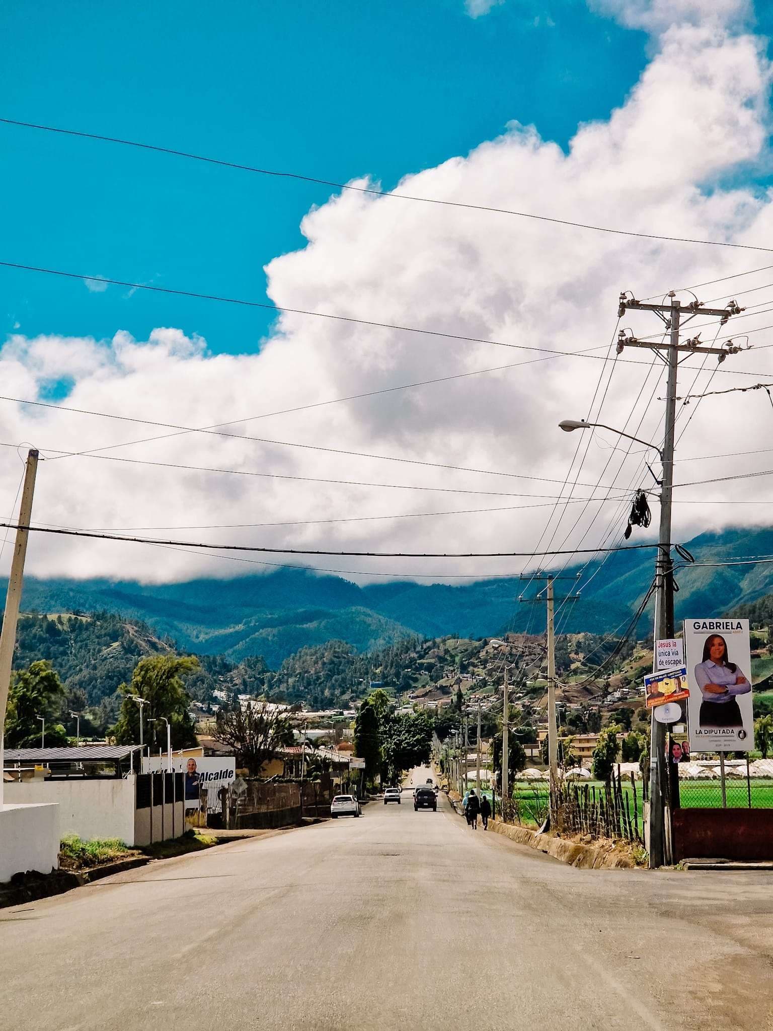 A paved road winds through a rural town with mountains in the background, featuring political campaign posters on utility poles under a blue sky.
