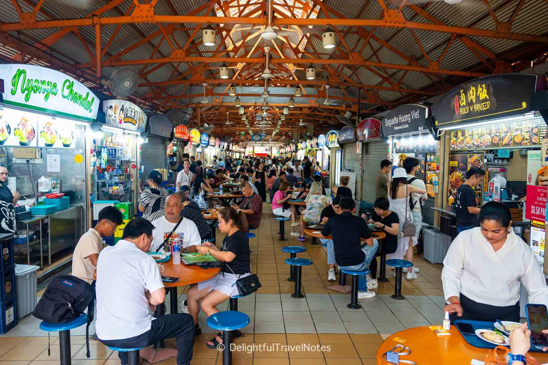Interior view of a busy hawker centre with orange steel roof trusses, food stalls, and diners eating at tables.