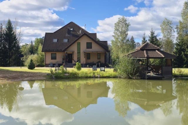 A beige house and a wooden gazebo sit on the grassy bank of a calm pond, reflecting in the water under a blue sky with scattered clouds.