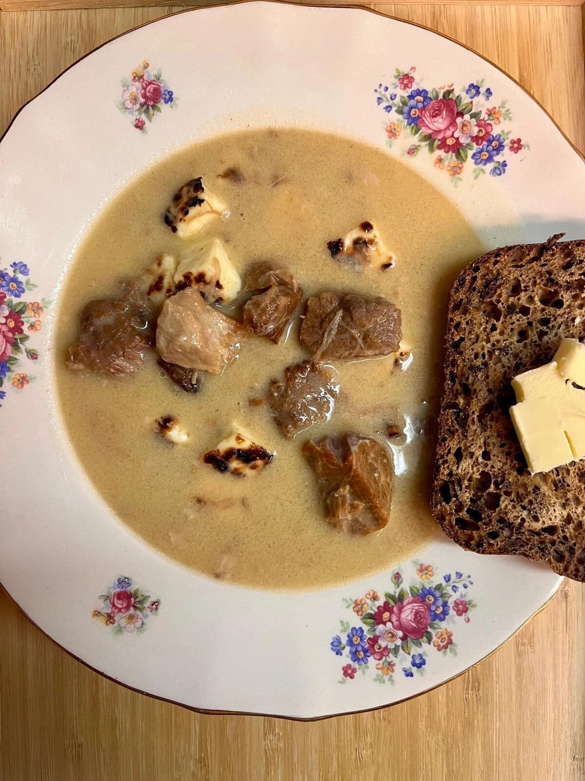 Overhead shot of a bowl of creamy Finnish cheese stew with meat chunks and bread on a floral plate.