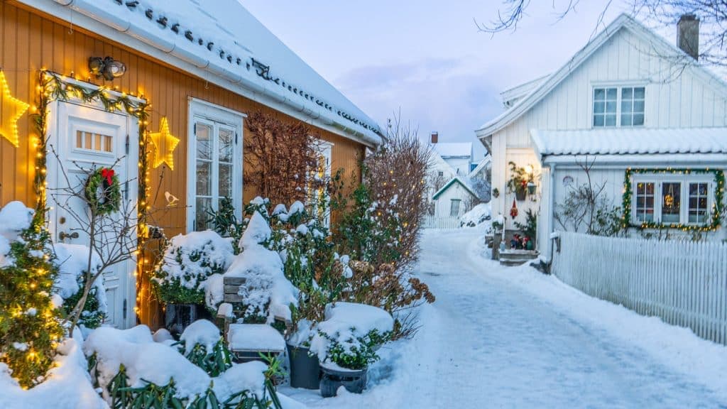 Snow-covered residential street in Drøbak featuring a yellow house with Christmas lights and a white house, capturing the festive winter atmosphere of Julehuset