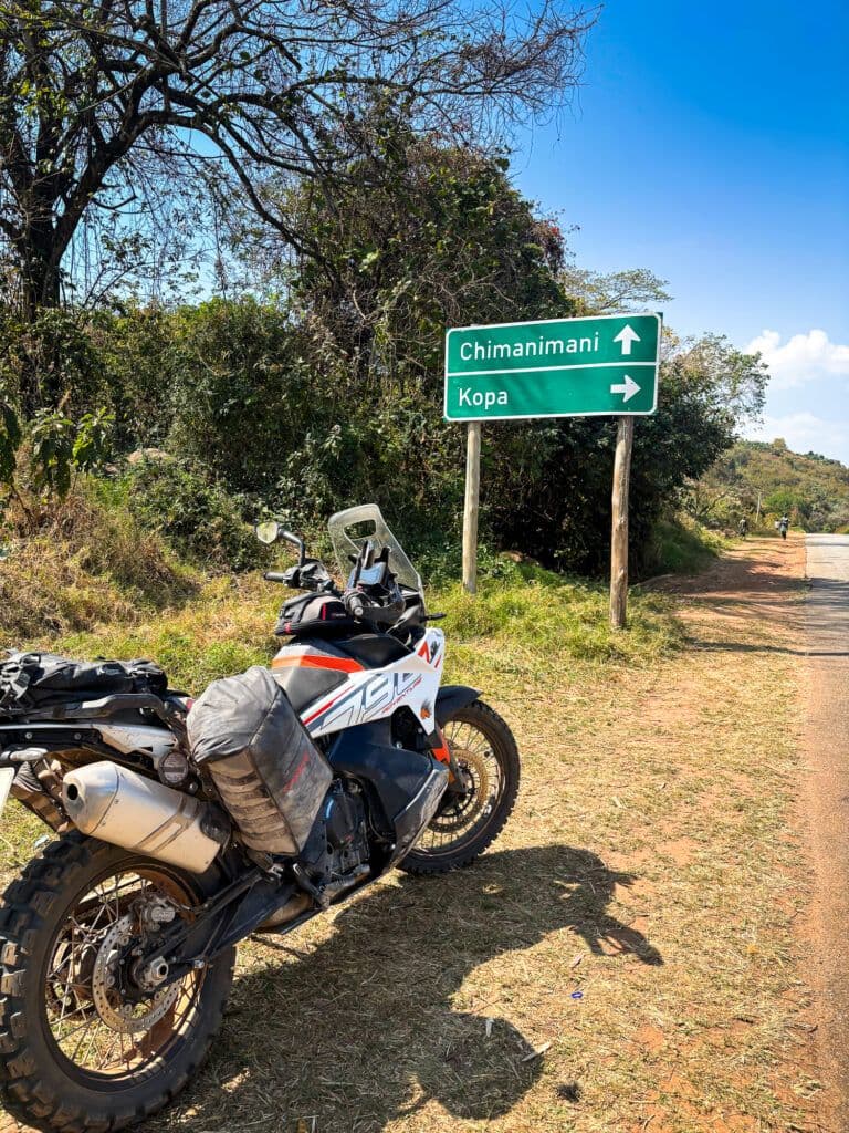 Packed adventure motorcycle parked on a dirt shoulder beside a green road sign pointing to Chimanimani and Kopa.