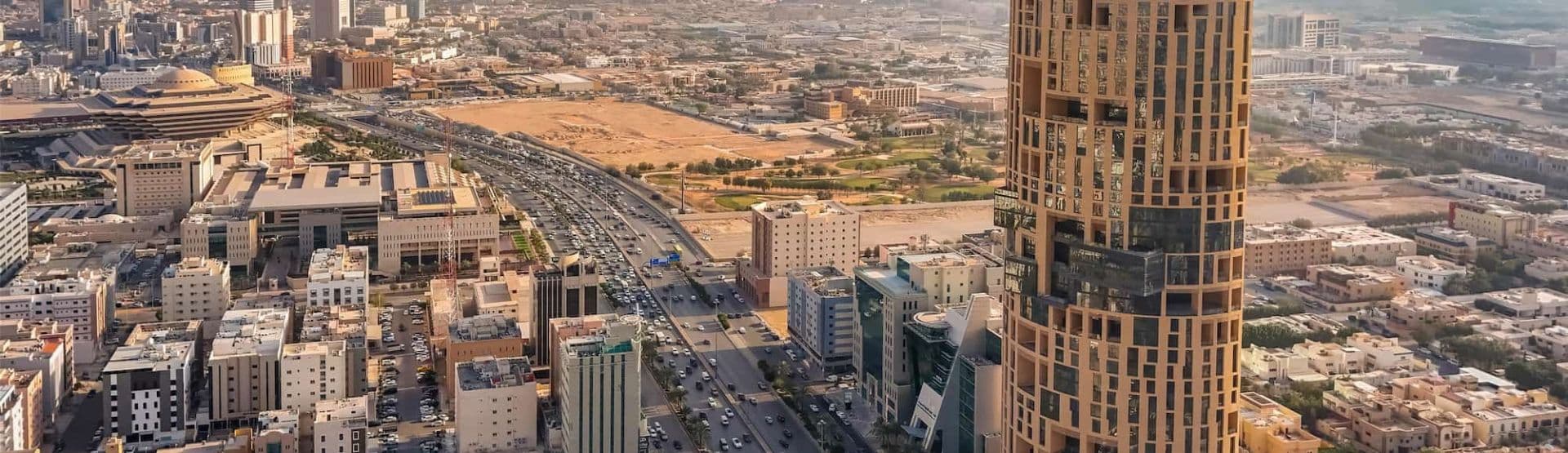 Aerial cityscape of the Kuwait region near Ash Shāmīyah showing a prominent skyscraper, highway traffic, and dense urban development under clear daylight