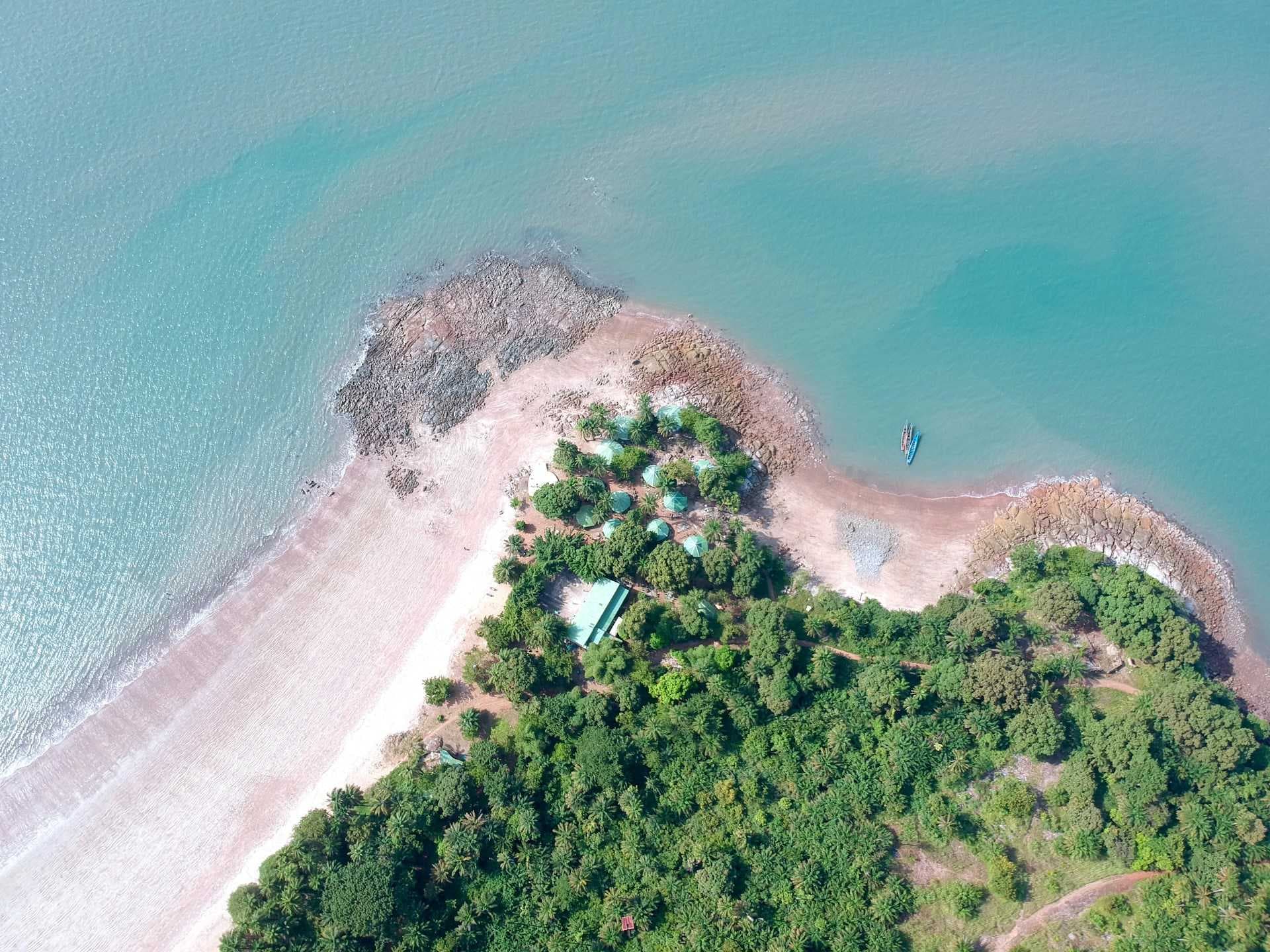 Aerial drone view of a coastal peninsula with turquoise water, sandy beach, rocky outcrops, green vegetation, and small boats.