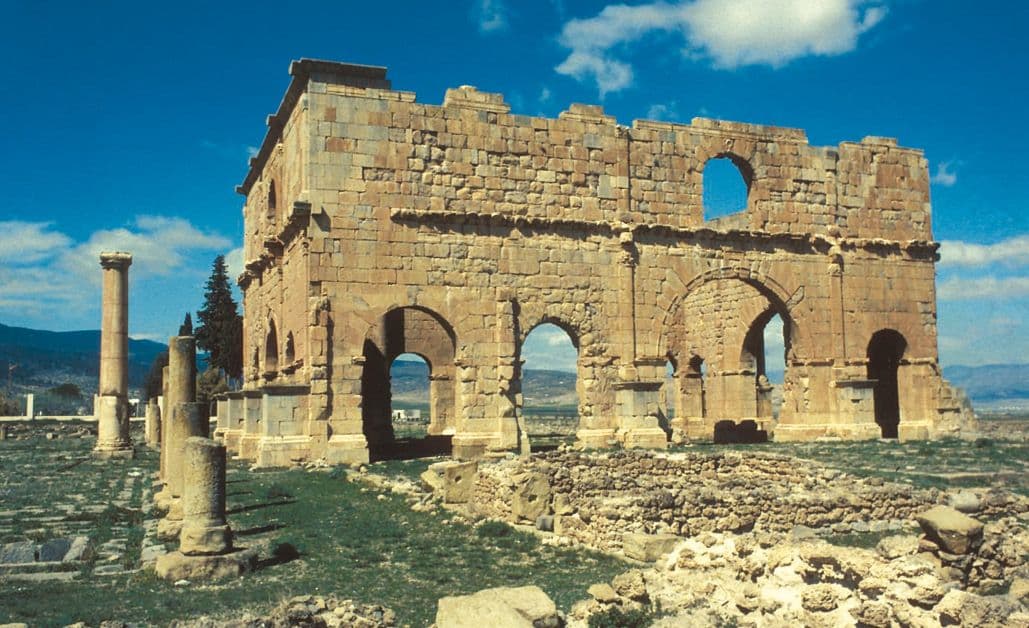 Ancient stone ruins featuring arched colonnades and standing pillars under a blue sky, depicting a classical archaeological site.