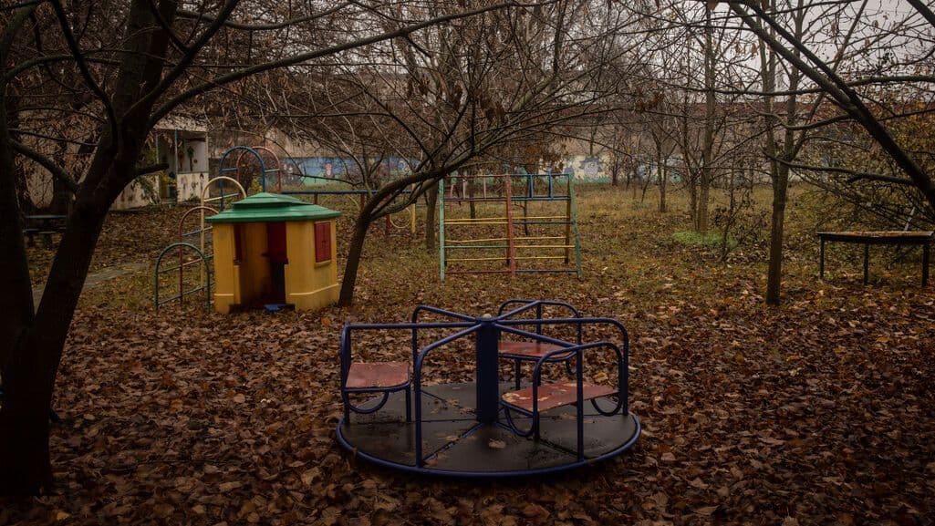 Autumn park playground with a blue merry-go-round, yellow playhouse, and climbing frame amid fallen leaves and bare trees.