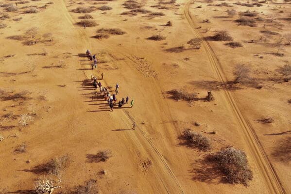 Aerial view of a group of people walking along a dusty, unpaved road through the arid, scrub-covered landscape of the Bakool region.