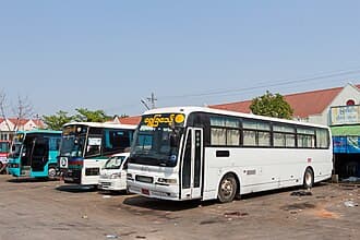 A white bus with yellow signage is parked in a dusty lot alongside other buses, likely at a transport hub in Pyapon.