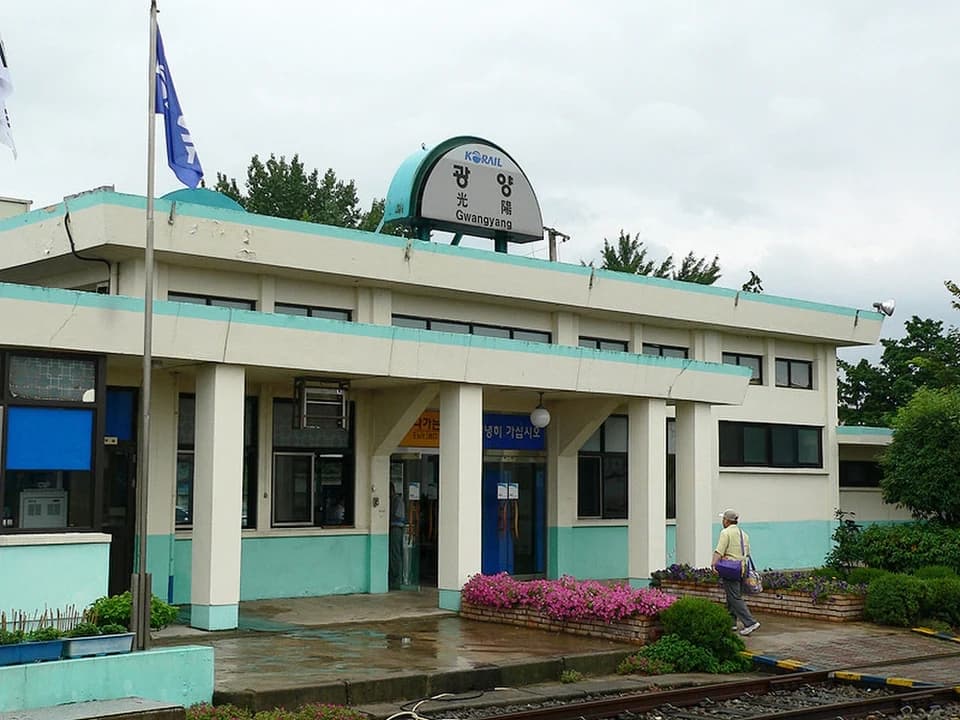Exterior of Gwangyang Station with KORAIL signage, teal and white building, flowers, and a person walking near the tracks