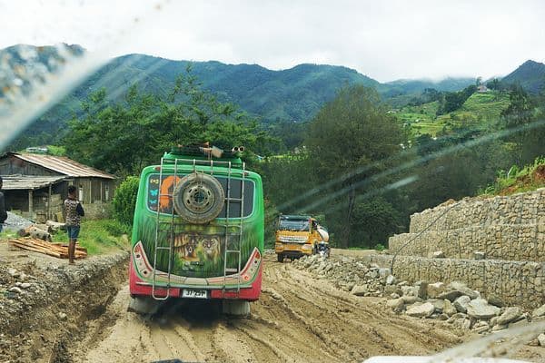 Decorated bus and truck navigating a muddy, unpaved mountain road through the Ermera interior, the typical route toward Laclo from Dili.
