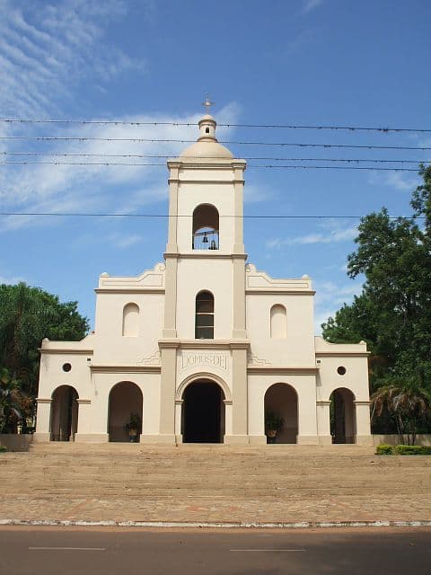 A cream-colored church with a bell tower and arched entrances stands on a raised stone platform under a blue sky.