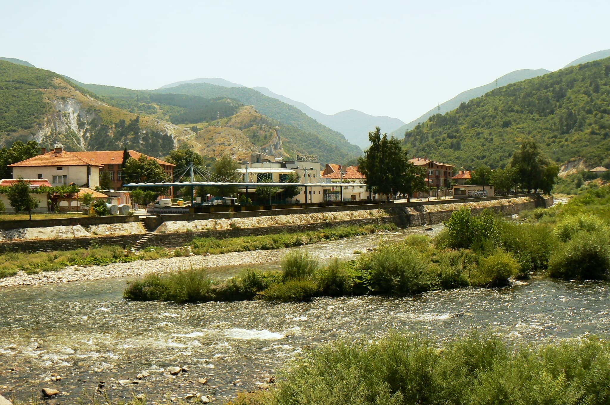Wide exterior view of Krichim town featuring a shallow river, stone embankment, buildings with red roofs, and green mountains in the background.