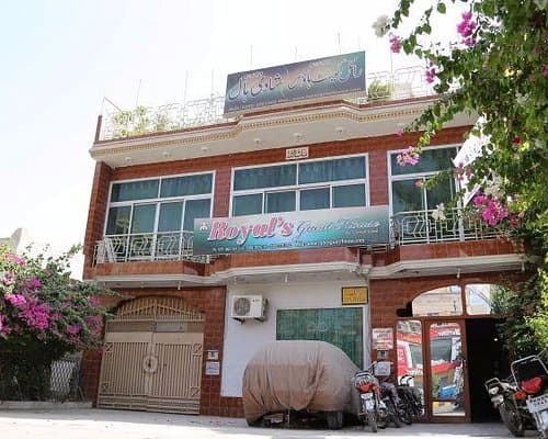 Exterior of a two-story brick building housing Royal's Guest House in Hyderabad, Sindh, featuring a covered vehicle and motorcycles parked on the street.