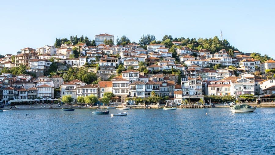 Lake Ohrid shoreline with waterfront buildings, boats, and hillside town under clear sky.