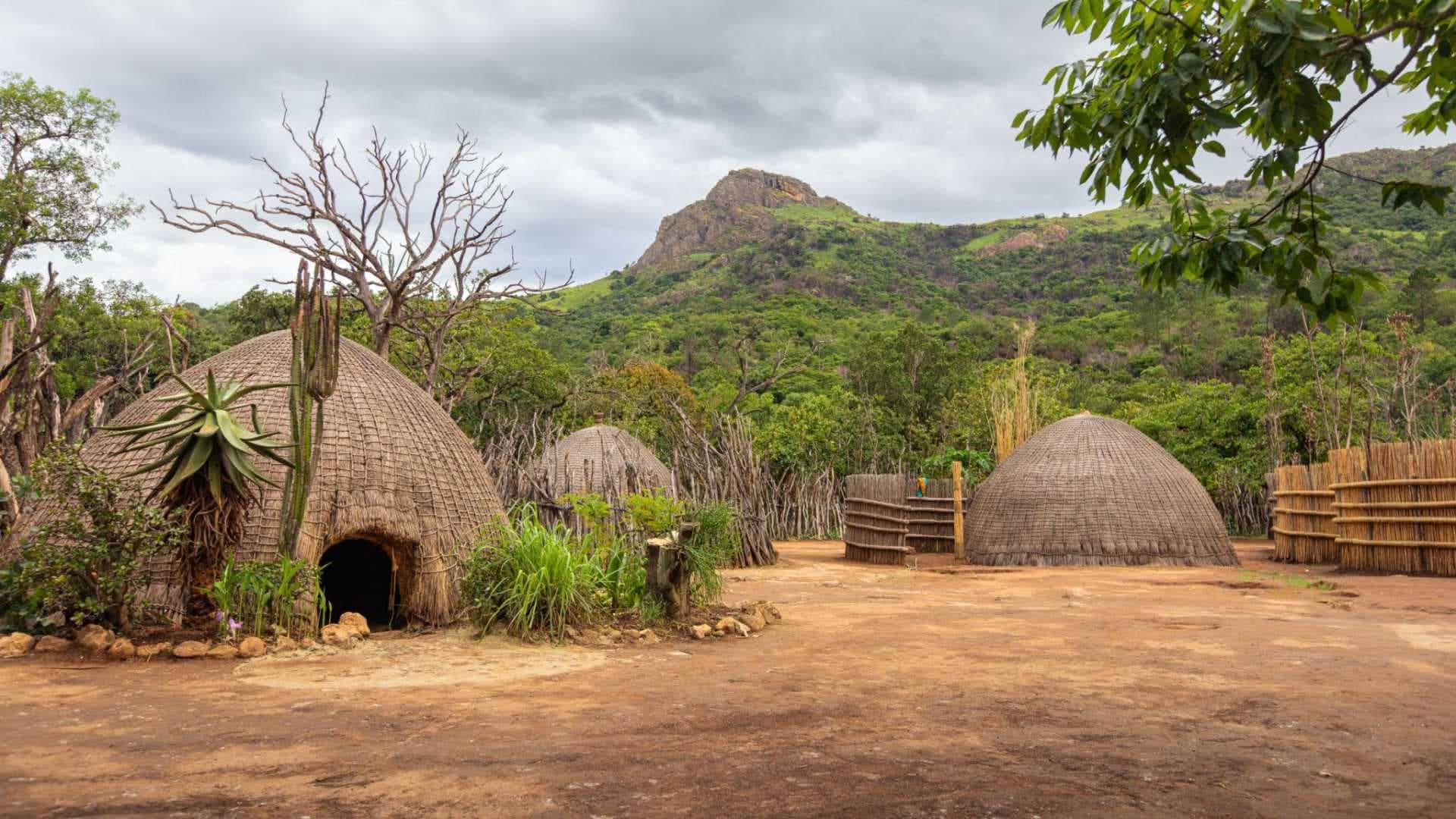 Traditional thatched rondavel huts in a rural Swazi homestead with green hills and overcast sky