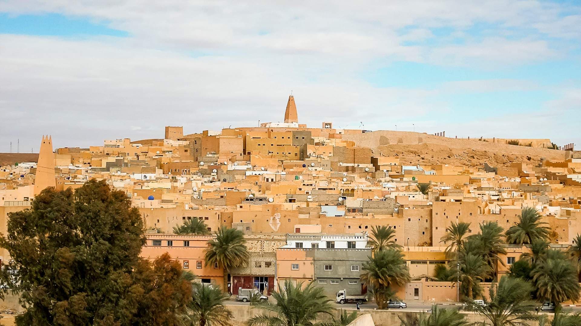 Wide view of Smala in the M'zab Valley, showing earthen buildings, minarets, palm trees, and dry terrain under a cloudy sky.
