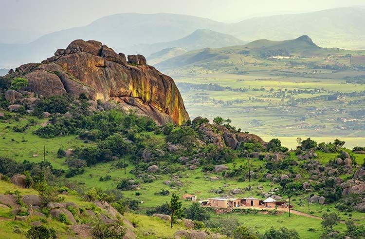 Green rolling hills and rocky outcrops under a hazy sky in the Eswatini countryside near Nsoko, with distant mountains and small rural structures.