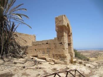 Sunlit stone ruins of a historical structure in Al Abyār, featuring a prominent arched tower and palm trees against a clear blue sky.