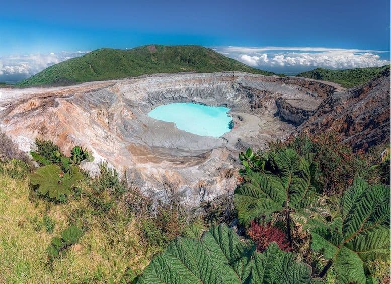 Aerial view of Poás Volcano's crater featuring a vibrant turquoise acidic lake surrounded by barren volcanic rock and lush green vegetation.
