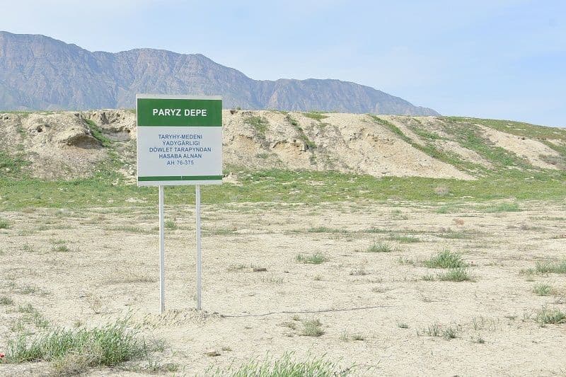 A green and white historical site sign for Paryz Depe stands in a dry, grassy field with hills in the background.