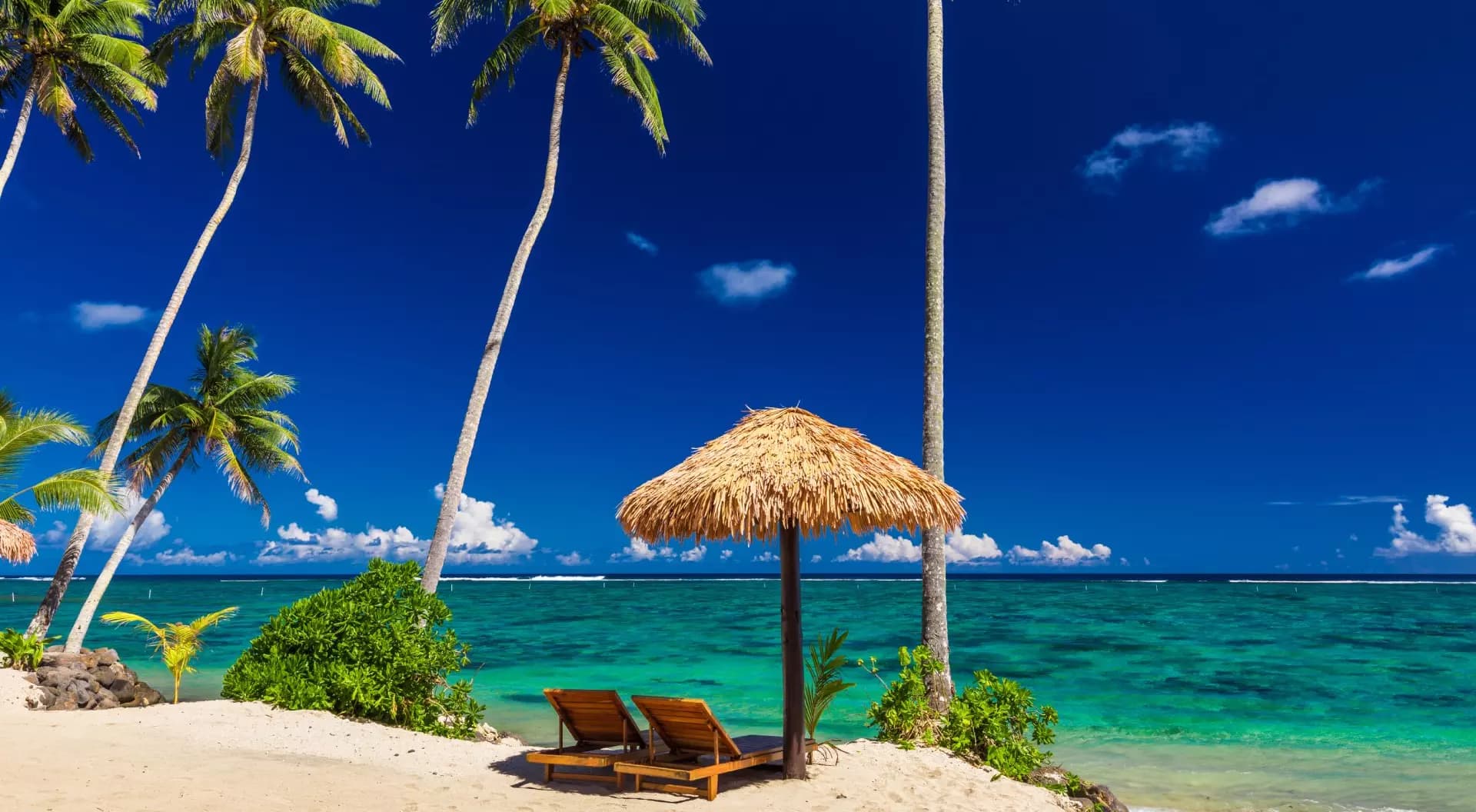 Tropical beach scene with palm trees, thatched umbrella, lounge chairs, turquoise water, and clear blue sky in Samoa.