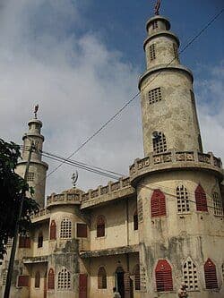 Exterior view of a historic town building with distinctive cylindrical towers, red-shuttered windows, and a crenellated roofline under a partly cloudy sky.