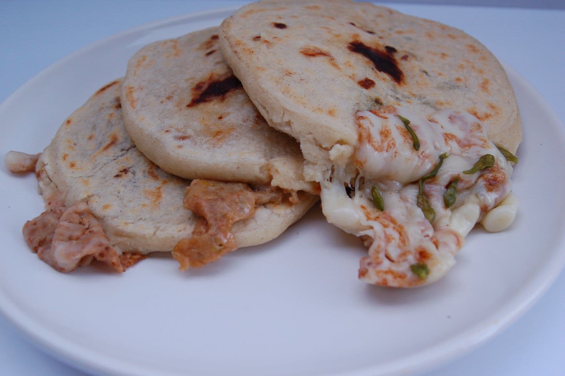Close-up photograph of three stuffed pupusas on a white plate, showing melted cheese and meat fillings spilling out.