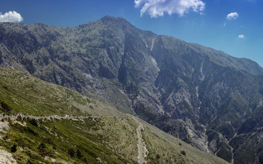 A winding mountain road descends through a rugged alpine valley toward distant peaks under a blue sky.