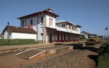 Exterior view of a historic railway station building with a yellow locomotive on the tracks under a clear blue sky.