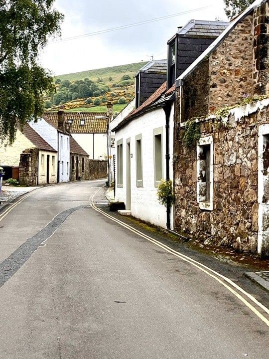 Quiet rural village road lined with stone cottages and rolling green hills, typical of the approach to Almondbank in Perthshire.