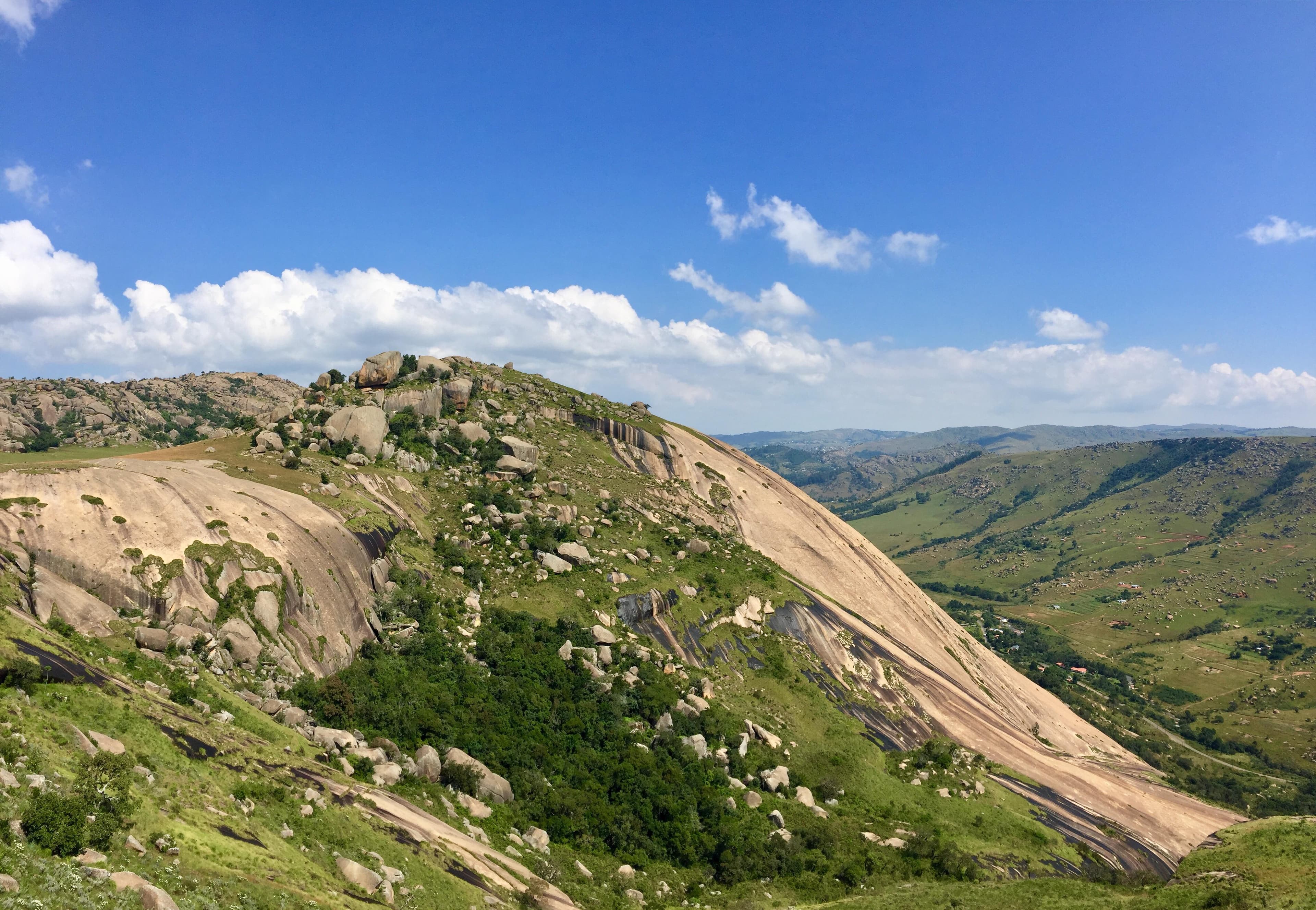 Wide exterior view of the massive Sibebe Rock granite peak, featuring smooth slopes, scattered boulders, and green vegetation under a blue sky.