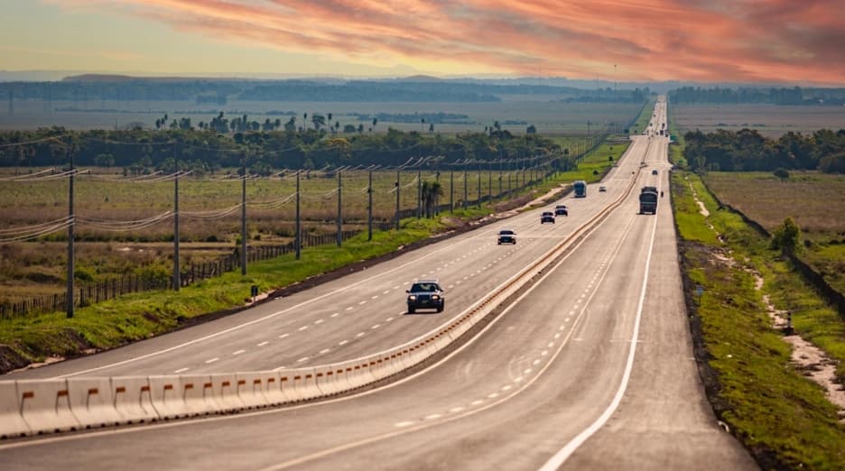 Wide highway stretching into the distance under a sunset sky, with vehicles, fields, and power lines.